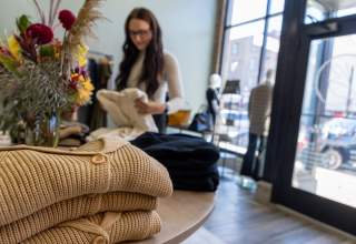 A young woman, slightly out of focus in the background, picks up a sweater in a well-lit boutique with huge windows. In the foreground are tan and navy blue cardigans on a table with a colorful flower arrangement in the middle of the table.