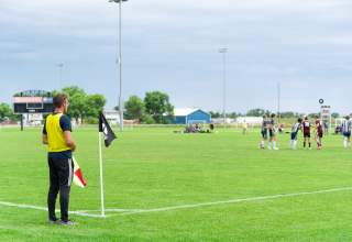Assistant referee standing at the corner of a green soccer field holding a flag, with players gathered in the distance near midfield under a cloudy sky and stadium lights.