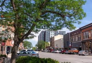 St. Joseph Street in downtown Rapid City with brick buildings, leafy trees and parked cars under a clear blue sky. The scene conveys a vibrant, sunny atmosphere.