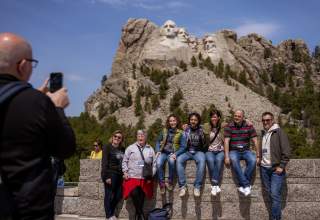 A group of seven people pose for a photo sitting on a stone ledge, looking at a cameraman on the edge of the photo to the left. Mount Rushmore stands in the background.