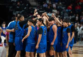 A basketball team in blue uniforms bring their hands together above their huddle at the end of  team timeout. Fans in the stands are visible out of focus in the background.