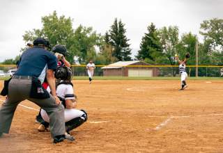 A softball pitcher in a white uniform fires a pitch toward home plate, where a batter in a blue uniform, the catcher, and umpire in navy blue await the pitch in the foreground. Mature trees stand just beyond the outfield fence under a cloudy sky.