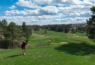 A man dressed in a black polo and black shorts tees off at the Golf Club at Red Rocks. Pine trees, a perfectly manicured course, bunkers, and a partly cloudy sky are visible in the distance.