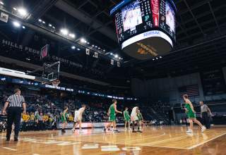 Viewed from court-level in the corner, two basketball teams are mid-game inside the Summit Arena. A large center-hung scoreboard displays game information for the fans visible in the stands.