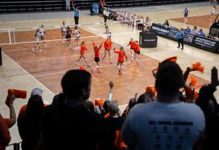 A volleyball team in orange uniforms cheer in focus in the background after scoring a point as supporting fans, out of focus in the foreground, clap.