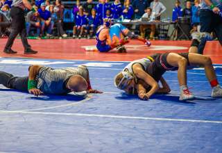 Wrestler attempting a pin on a blue mat during a match as a referee lies close to the action to check the shoulders, with another match and spectators visible in the background.