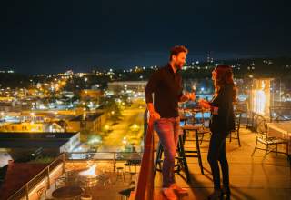 Couple clinking glasses in the evening surround by city lights on the roof of Vertex Sky Bar at Hotel Alex Johnson in Downtown Rapid City, SD