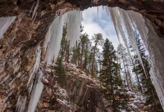 View from inside a rocky ice cave with large icicles hanging from the ceiling. Snow-dusted evergreen trees are visible outside against a cloudy sky.