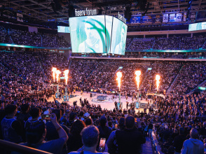 A packed crowd fills Fiserv Forum in Milwaukee during an NBA game as flames shoot up from the court during player introductions, with the arena’s large video scoreboard displaying a close-up of a player.