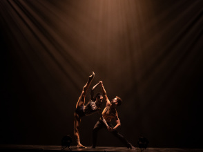 Two contemporary dancers perform an expressive lift under dramatic stage lighting.