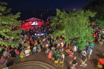 Dozens of people watching a concert at Memorial Union Terrace at night. The photo is taken from above showing the stage, people, and the popular yellow, orange, and green terrace sunburst chairs.