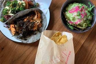 A plate of tacos with dark gray tortillas, a small bowl of guacamole with a bright pink garnish, and a small bag of tortilla chips lay on a table at Bandit