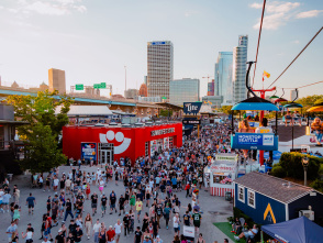 A large crowd moves through the bustling Summerfest grounds in Milwaukee during the day, with the downtown skyline visible in the background. A red building with the Summerfest smiley-face logo serves as the festival store. Above, colorful gondolas on the Skyglider transport people across the venue. The scene is filled with booths, food stands, and signage under a clear sky.