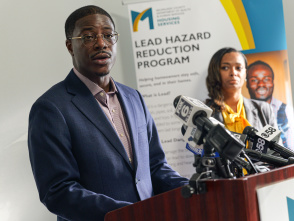 A man in a suit speaks at a podium during a press conference, with microphones from local media outlets in front of him and a banner about a lead hazard reduction program in the background.