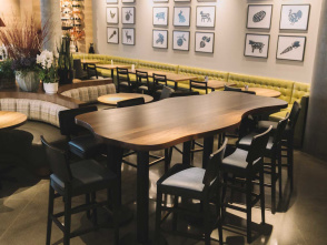 Modern dining area with a long wooden communal table surrounded by black chairs, cozy booth seating, potted plants, and framed artwork on the wall.