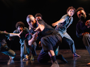 A group of six dancers strike expressive, dynamic poses on stage under dramatic lighting, wearing modern, casual performance outfits.