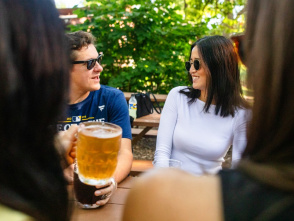Friends gather at an outdoor patio table surrounded by greenery, enjoying drinks and conversation on a sunny day. A glass of beer is raised in the foreground while two people wearing sunglasses smile and chat, creating a relaxed and social atmosphere.