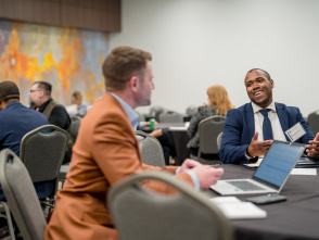 Two business professionals conversing at a conference table - one in an orange blazer with back to camera, the other in a navy suit smiling and gesturing while speaking. Multiple attendees visible in background conference room setting.