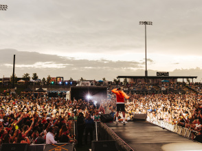 A performer in a red hoodie stands on an outdoor stage facing a massive crowd at sunset. Thousands of fans pack the venue, many holding up phones to capture the moment, while bright stadium lights and food stand signs glow in the background.