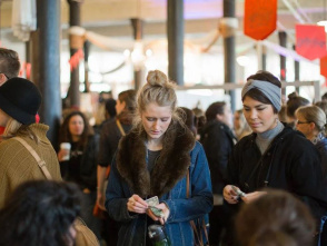 A busy indoor market scene with people browsing and making purchases. Two women in the foreground count money, while crowds of shoppers fill the background among vendor booths and hanging signs.