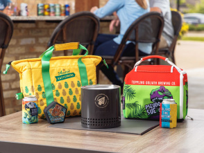 Colorful drink coolers and canned beverages displayed on an outdoor table, including a yellow pineapple-patterned cooler, a red-and-green cooler with a dinosaur graphic, and a compact black tabletop fire pit.