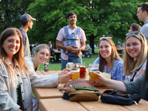 A group of friends sitting at a park picnic table smiling at the camera as they raise their glasses to cheers, with a group of men talking behind them, with trees in the background.