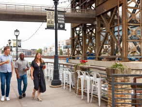 Three people walk along the Milwaukee Riverwalk outside Lakefront Brewery, holding beers and smiling. They pass under an industrial bridge structure with banners reading “Cold Beer in Here” and “Beer. Food. Beer.” while white bar stools and flower planters line the railing overlooking the river.