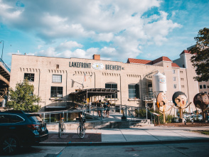 Exterior view of Lakefront Brewery in Milwaukee, a historic brick building with large windows, outdoor entrance ramp, and people gathered at the front. A white brewing tank and whimsical oversized face sculptures are visible on the right, with cars and bicycles in the foreground under a partly cloudy sky.