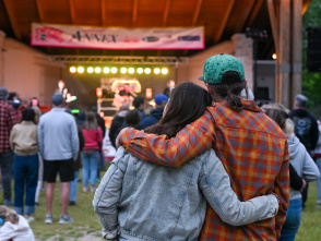 A couple stands arm in arm watching a live outdoor concert from the audience. Stage lights glow beneath a covered pavilion as community members gather on the lawn to enjoy the performance.