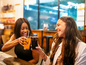 Two women smiling and clinking glasses of Sprecher soda at a wooden table inside a brewery