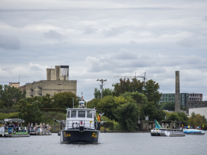 A river scene featuring three boats on calm water; a ferry with passengers, another small boat, and an industrial backdrop of factories and greenery. Cloudy sky.
