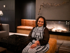 Annette Jackson wearing a black and white houndstooth dress with black cardigan sits smiling in an upscale modern lounge. She is positioned on a tan leather chair in front of a contemporary fireplace with a dark stone surround. The space features modern interior design with neutral tones, additional seating visible in the background, and warm lighting that creates an elegant, sophisticated atmosphere.
