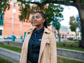 Ashley Jordan stands outdoors in an urban setting, wearing a tan trench coat over a dark shirt, with braided hair and a slight smile, with brick buildings and trees visible in the background