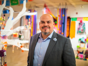Jacobo Lovo of Latino Arts stands smiling in a colorful community space, wearing a gray blazer and light blue shirt with a decorative pin