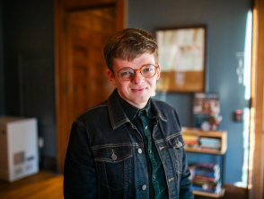 Ryan Castelaz with short brown hair and round glasses smiles warmly at the camera. They're wearing a dark denim jacket over a dark green plaid shirt, standing in what appears to be a cozy indoor space with warm wood tones, bookshelves, and soft lighting in the background.