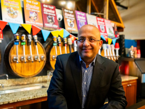 Sharad Chadha, CEO of Sprecher Brewing Company, sits smiling in a dark suit and blue checkered shirt at the brewery's taproom bar. Behind him are multiple beer taps, colorful triangular bunting, and shelves displaying various Sprecher product bottles and signage.