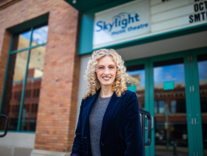 Susan Varela, executive director of Skylight Music Theatre, smiles while standing in front of the theater's brick building with the Skylight music theatre sign visible in the background