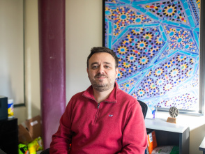 Milwaukee Rep Director Chad Bauman wears a red shirt and sits with hands clasped in his office with geometric artwork in the background.