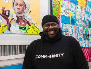 a portrait of a black man laughing in front of a gallery wall of colorful art with a black beanie and black hoodie that reads "COMMUNITY" on it in white lettering