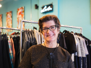 a portrait of a white woman with cat eye glasses smiling in front of a rack of clothes