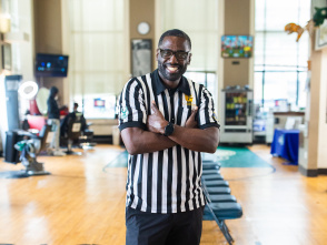 Gaulien 'Gee' Smith, a Black man wearing glasses and a black and white striped referee shirt, stands with his arms crossed and a big smile in what appears to be a gym or recreation center. The polished wooden floor and exercise equipment are visible in the background, along with large windows and colorful wall decorations.