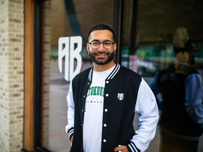 Nimesh "Nemo" Ruparel stands smiling outside a storefront wearing a black and white varsity-style jacket with "MILWAUKEE" text in green letters and "AB" logo. He has glasses, a beard, and is positioned in front of glass windows with "AB" lettering visible in the background. The setting appears to be an urban commercial area with brick architecture.