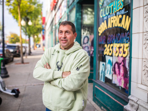 Peter Feigin in a light green hoodie with a deer logo stands with his arms crossed, smiling at the camera on a city sidewalk. Behind him is a colorful storefront window with "CLOTHES" and other text visible, along with decorative stone architecture. Trees and parked cars line the urban street scene.