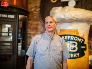 Russ Klisch wears a light blue button-down shirt at Lakefront Brewery. Behind him are exposed brick walls, industrial-style lighting, and a large format frothing beer mug.
