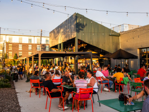 A large patio at a brewery is full of people sitting in red chairs. The golden light of sunset spills over the space. A large bar is in the background with a green overhang.