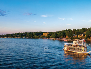A tour boat cruises across a calm lake at sunset, with passengers aboard and shoreline homes nestled among trees in the background. Warm golden light reflects off the water, creating a scenic and relaxing lakeside view.