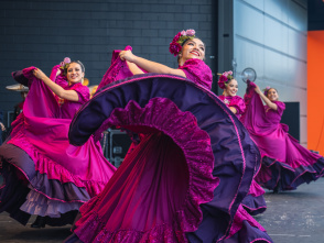A group of female dancers performs a traditional Mexican folklórico dance on stage. They wear vibrant magenta dresses with purple ruffles and floral headpieces. The lead dancer in the foreground smiles brightly while gracefully twirling her flowing skirt. The dancers behind her mirror the movement, creating a colorful, energetic scene against a modern stage backdrop.