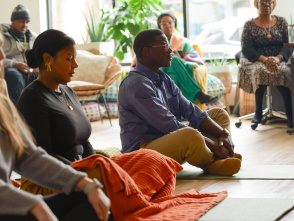 A diverse group of adults sit on the floor and in chairs during a small group discussion or community gathering in a bright, welcoming indoor space.