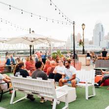 A group of people enjoy outside at Bridgeview Box Park. The Cincy skyline is in the background.