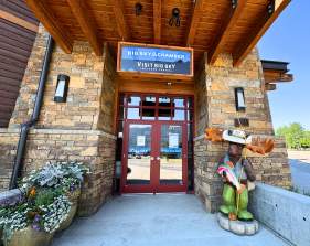 Big Sky Chamber/Visit Big Sky entrance with stone facade, red doors, floral planters, and a green moose statue with fishing gear.